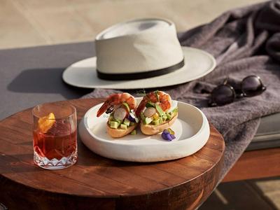 Due panini con farcitura su un piatto bianco con bevanda, cappello e occhiali da sole su tavolo in legno.