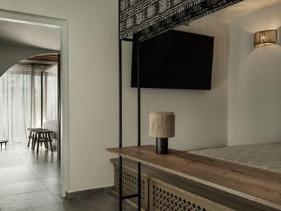 Área moderna de habitación de hotel con televisor, lámpara de mesa y vista a una habitación luminosa con mesa de comedor.