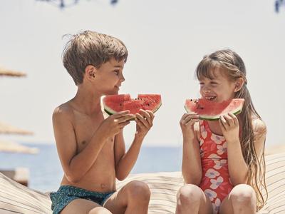 Dos niños sentados en la playa comiendo sandía bajo el sol.