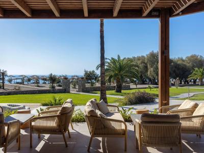 Terraza cubierta con sillas y vista a jardines y palmeras junto al mar bajo un cielo azul claro.