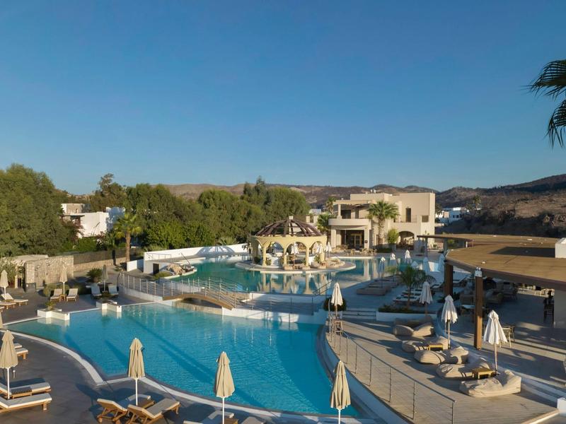 Gran área de piscina al aire libre de un hotel con tumbonas, sombrillas y panorama rodeado de palmeras.