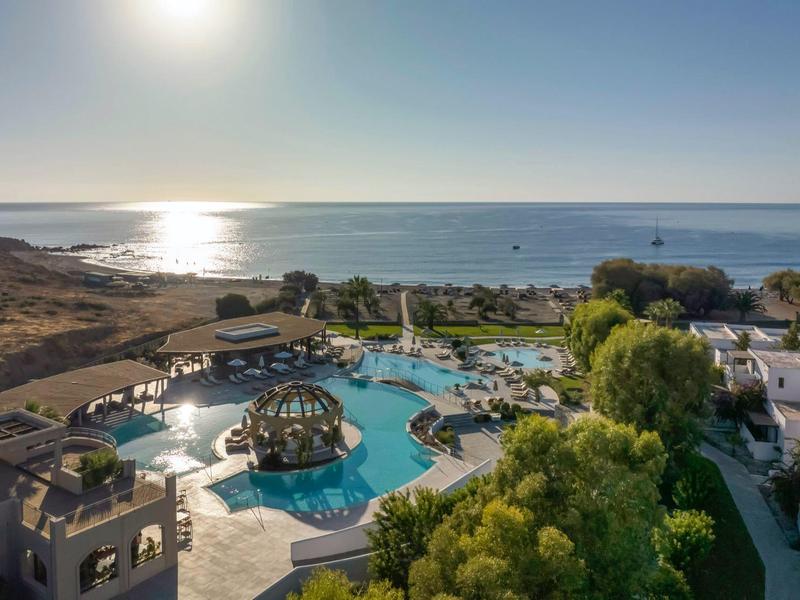 Vista de un resort con piscina, juegos de agua y jardín cerca del mar con luz solar.
