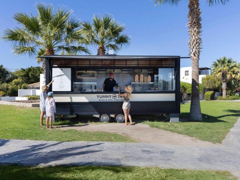 Quiosco moderno con bebidas y comida al aire libre, rodeado de palmeras y áreas verdes.