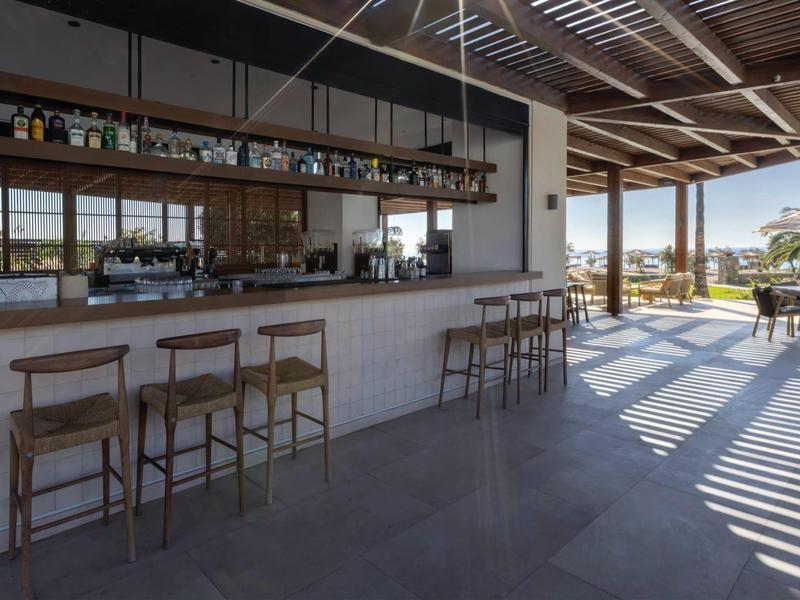 Área moderna de bar de hotel con taburetes de madera y grandes ventanales con vista al exterior.