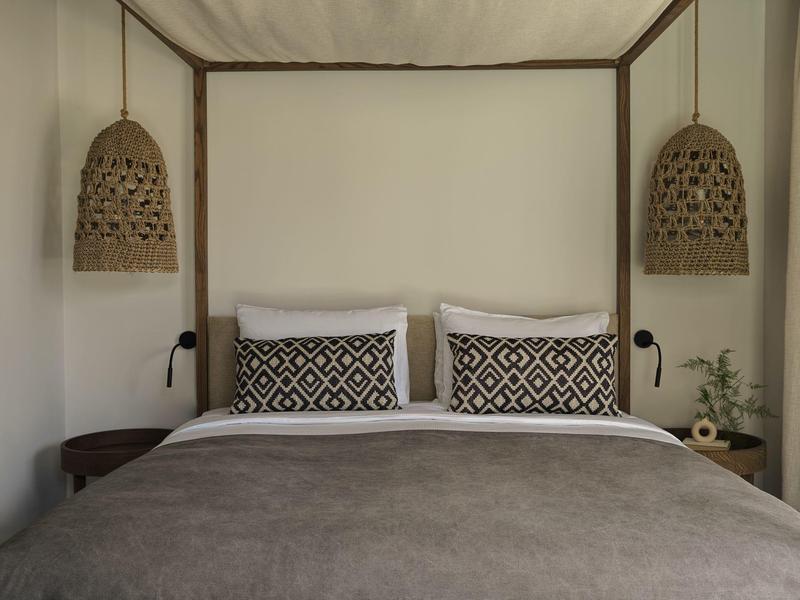 Cama doble sencilla con manta marrón, cojines con patrón y dos lámparas colgantes de mimbre en la pared.