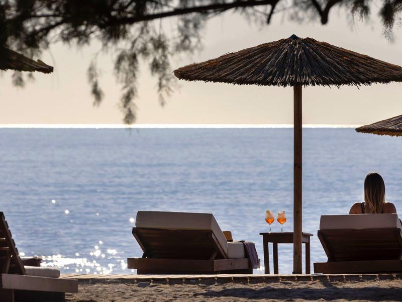 Una persona está sentada bajo una sombrilla en la playa mirando el mar brillante.