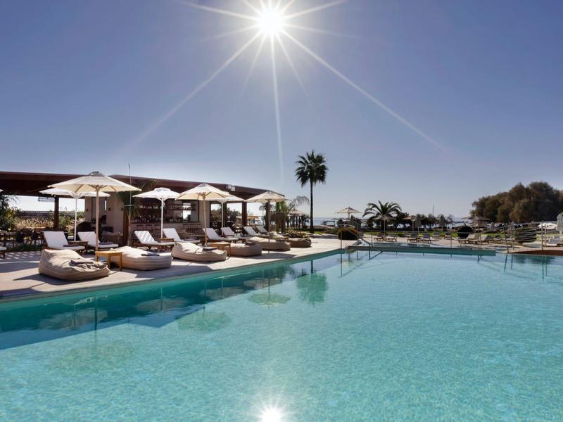 Piscina exterior luminosa con tumbonas y sombrillas bajo un cielo azul claro.