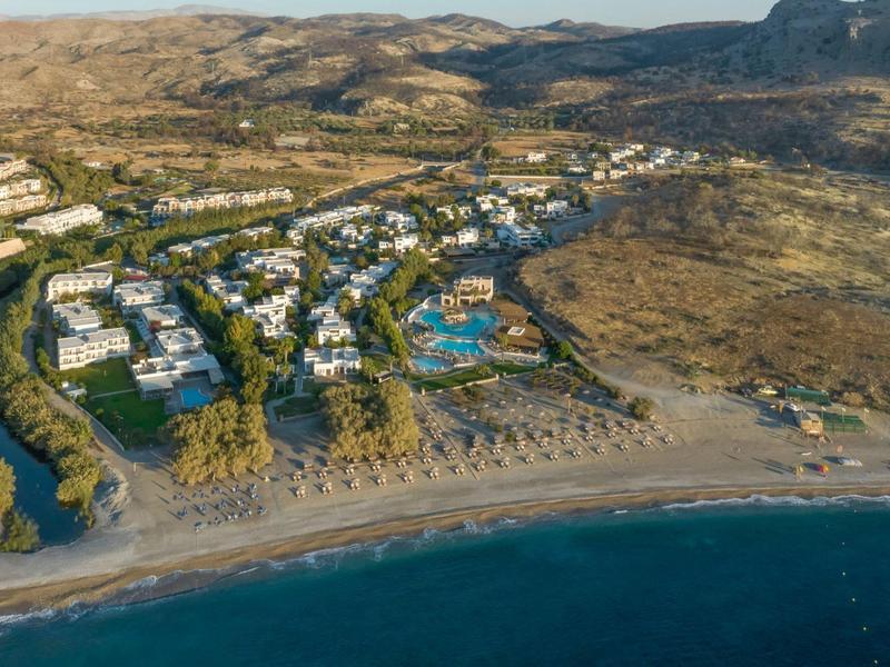 Vista aérea de un hotel con piscina junto a la playa y montañas cercanas.