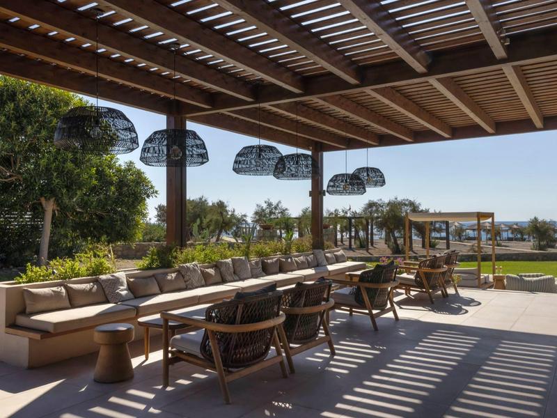 Terraza abierta con sillas de madera y sofás bajo un techo de madera con vista al jardín y al mar.