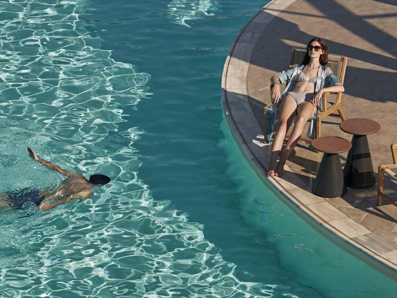 Mujer relajándose en una tumbona al borde de la piscina mientras una persona nada en la piscina.