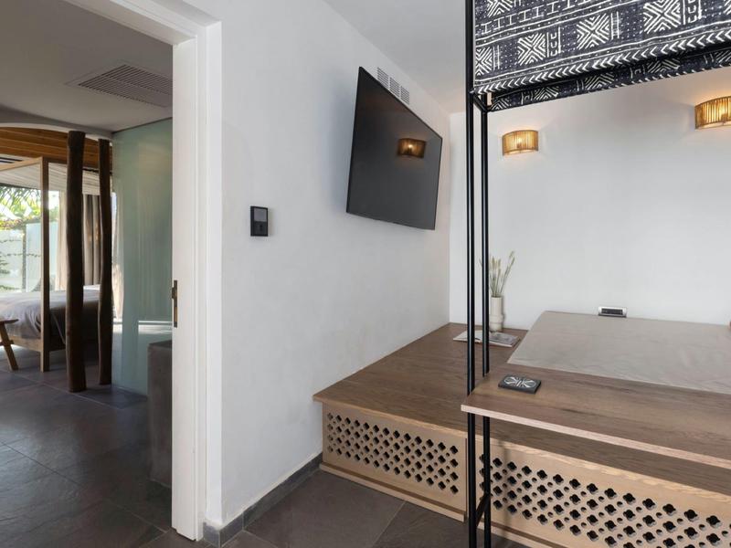 Área moderna de habitación de hotel con televisor de pared, banco y vista a la entrada con fachada de vidrio.