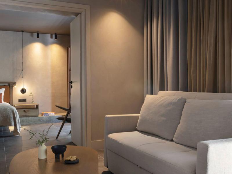 Sala de estar con sofá blanco y mesa, conectada a un dormitorio con cama y lámparas colgantes.