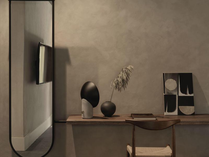 Interior moderno de habitación de hotel con espejo redondo, televisor, esculturas y estante de madera en pared gris.