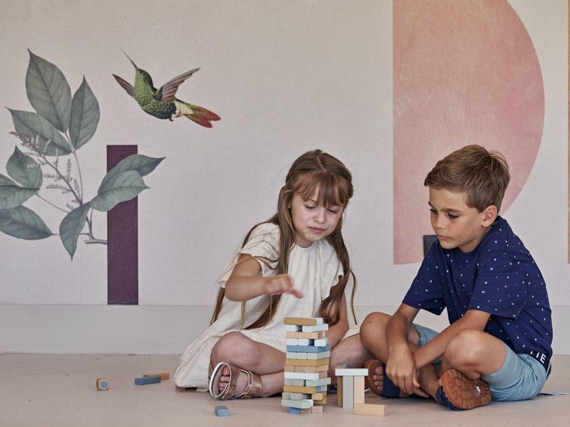 Dos niños juegan juntos a un juego de bloques de madera frente a una pared decorada con motivos de aves y plantas.