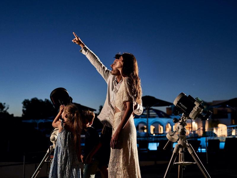 Tres personas miran al cielo estrellado de noche con un telescopio, una señala hacia arriba.