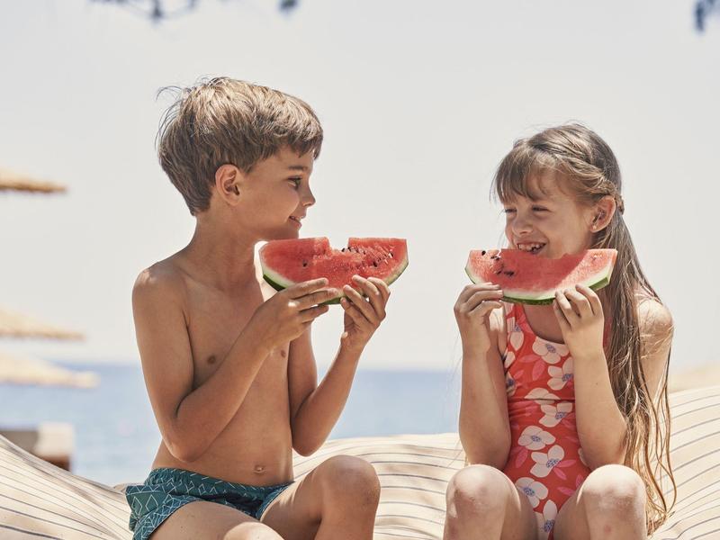 Dos niños sentados en la playa comiendo sandía bajo el sol.