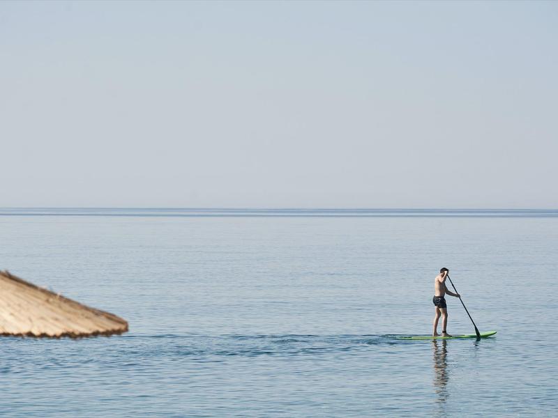 Persona está de pie en una tabla de paddle en el mar tranquilo frente a una pequeña sección de arena.
