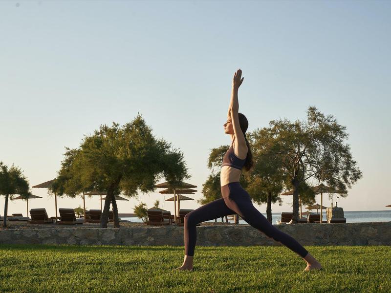 Mujer practica yoga al aire libre sobre césped verde con árboles y tumbonas de fondo.