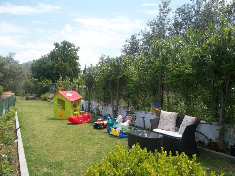 Garten mit Rasen, Kinderspielzeug, gelbem Spielhaus, Büschen und einer schwarzen Sitzgarnitur.
