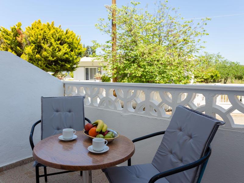 Balkon mit rundem Holztisch, zwei grauen Stühlen, Kaffeetassen und Obstschale bei klarem Himmel.