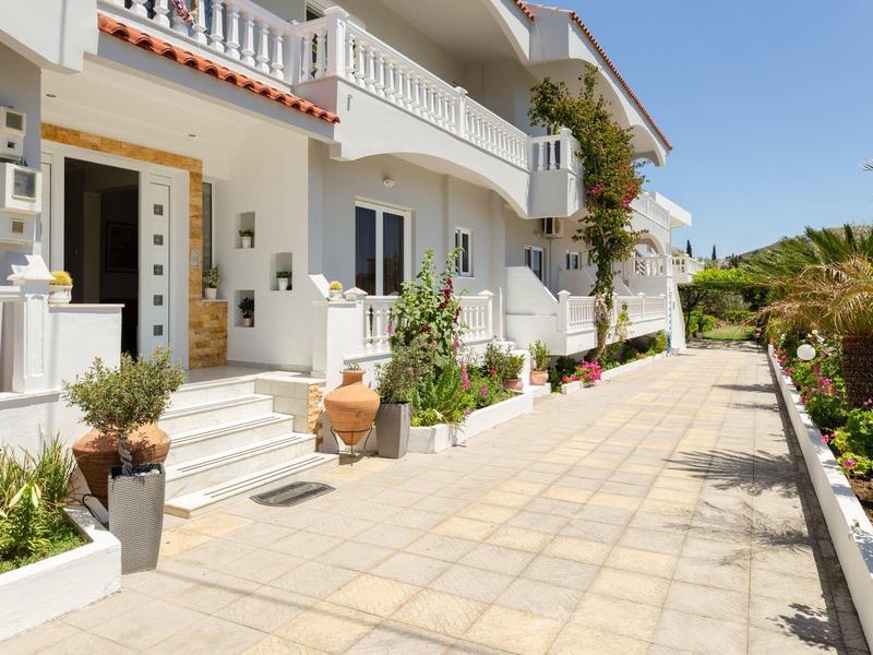 Weißes Wohnhaus mit Terrasse, Pflanzen in Töpfen und gepflastertem Weg bei sonnigem Himmel.