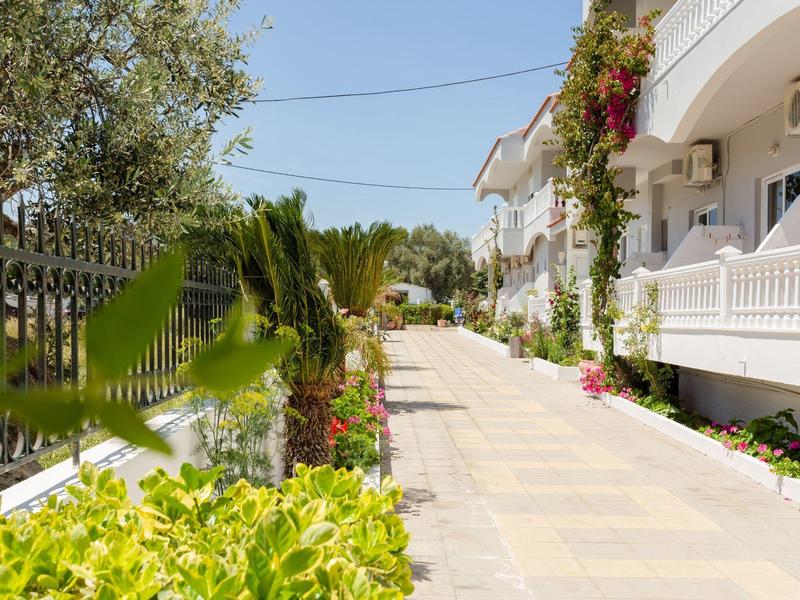 Schmaler Gehweg zwischen weißem Wohnhaus mit Balkon und üppigem Grün unter blauem Himmel.