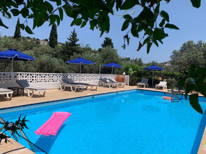 Blauer Pool mit Liegestühlen, Sonnenschirmen und grünen Bäumen im Hintergrund bei klarem Himmel.