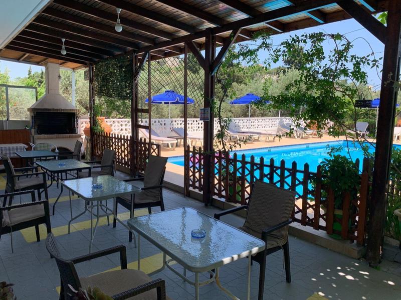 Überdachte Terrasse mit Tischen, Stühlen und Blick auf einen Pool mit blauen Sonnenschirmen.