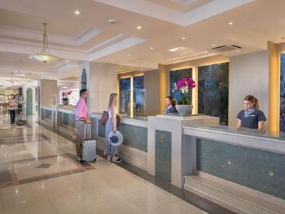 Reception dell'hotel con ospiti e personale, interno luminoso con bagagli da viaggio.