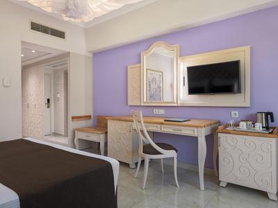 Hotelzimmer mit Fliederwand, Schreibtisch, Stuhl, Spiegel, TV und Bett mit braunem Überwurf.