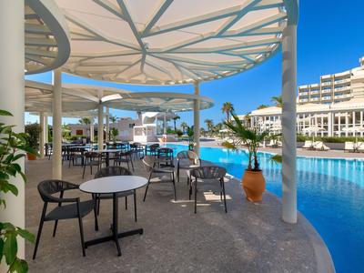 Area salotto con tavoli e sedie accanto a una piscina di un hotel in una giornata di sole.