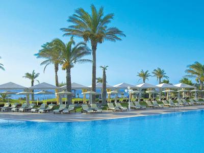 Grande piscina con palme e lettini sotto un cielo azzurro in un resort.