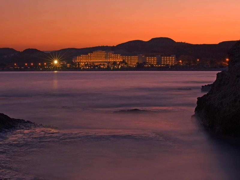 Sonnenuntergang über einer ruhigen Bucht mit Hotellichtern und Bergen im Hintergrund.