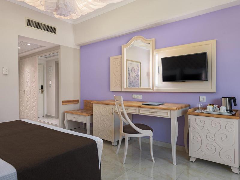 Hotelzimmer mit Fliederwand, Schreibtisch, Stuhl, Spiegel, TV und Bett mit braunem Überwurf.