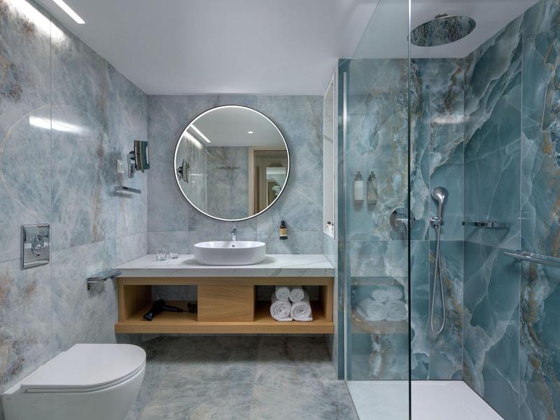 Modernes Badezimmer mit Marmorfliesen, rundem Spiegel, Waschtisch und Dusche mit Glaswand.