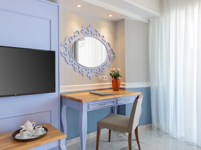 Helles Zimmer mit blauem Schreibtisch, grauem Stuhl, Spiegel und TV an der Wand.