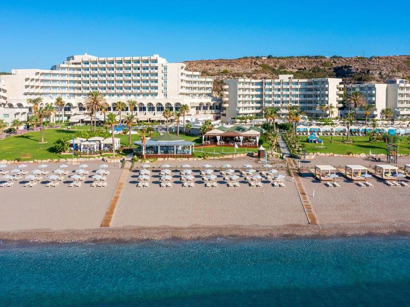 Großes Hotel mit Strand, Liegestühlen und klar blauem Wasser vor einem Hügel.