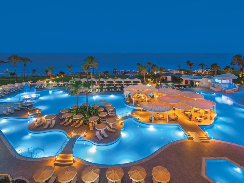 Großer, beleuchteter Hotelpoolbereich mit Palmen und Sitzgelegenheiten bei Nacht.