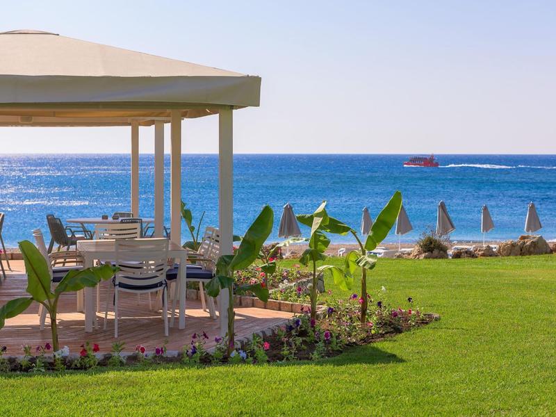 Terrasse mit Tischen und Stühlen am Meer mit Sonnenschirmen und Boot im Hintergrund.