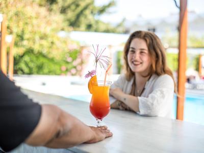 Ein bunter Cocktail auf einer Bar, im Hintergrund lächelt eine Frau am Pool.