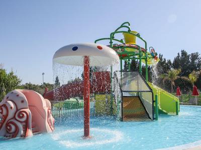 Bunter Wasserspielplatz mit Pilz-Dusche und Rutsche in einem flachen Pool unter blauem Himmel.