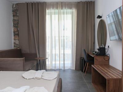 Helles Hotelzimmer mit Bett, Sofa, Schreibtisch, TV und bodentiefem Fenster mit Vorhängen.