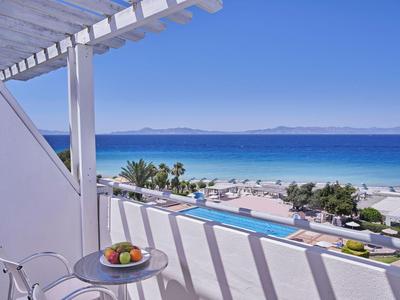 Balkon mit Tisch und Stühlen bietet Blick auf Strand, Meer und Pool unter klarem Himmel.