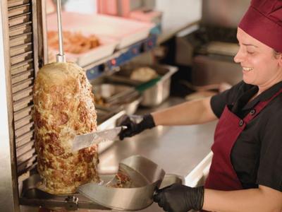 Eine Person in Küchenkleidung schneidet Fleisch vom Dönergrill in einer Restaurantküche.