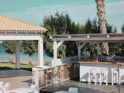 Terrasse mit Bar und weißen Stühlen mit Blick auf Palmen und das Meer im Hintergrund.