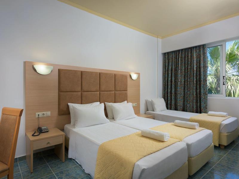 Bright hotel room with twin beds, yellow accents, wooden furniture, and a window showing palm trees.
