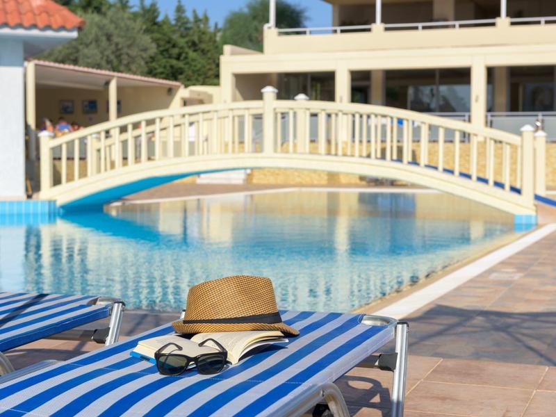 Sunny poolside with blue-striped loungers, a hat, and sunglasses by a small arched bridge.