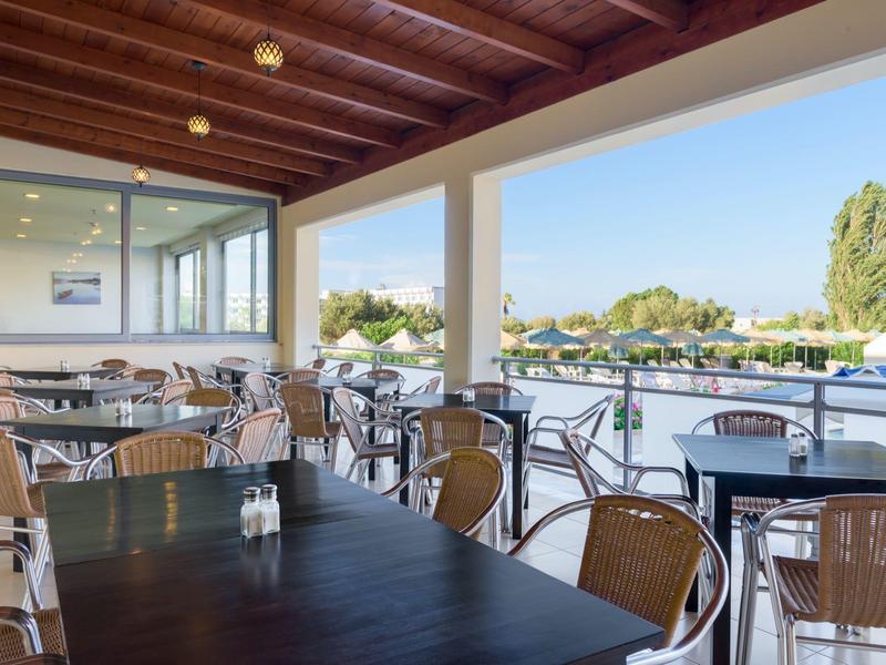 Covered patio restaurant with wooden tables and metal chairs overlooking a swimming pool and garden.