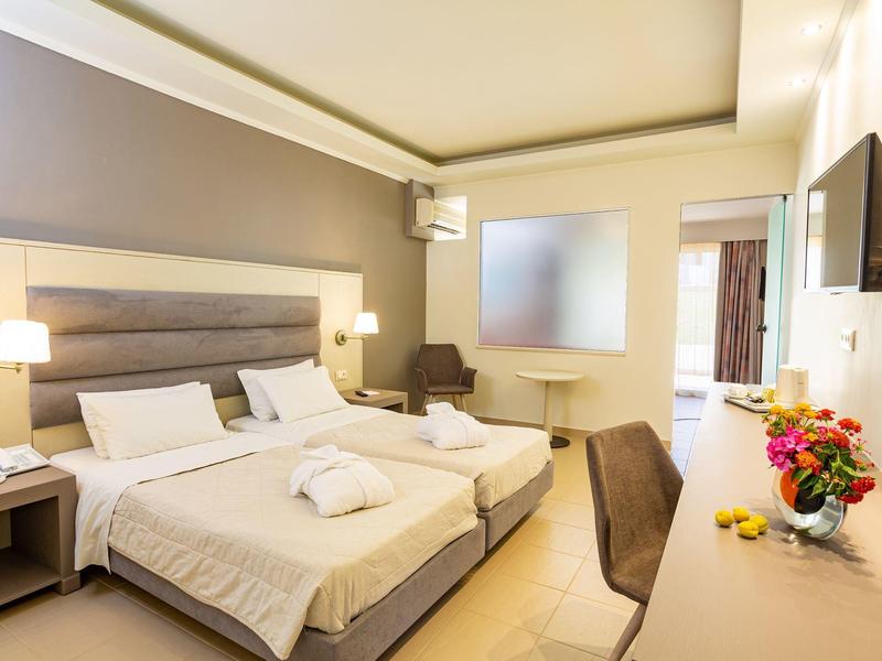 Jasny pokój hotelowy z dwoma łóżkami, stolikami nocnymi, krzesłem, biurkiem i telewizorem na ścianie.