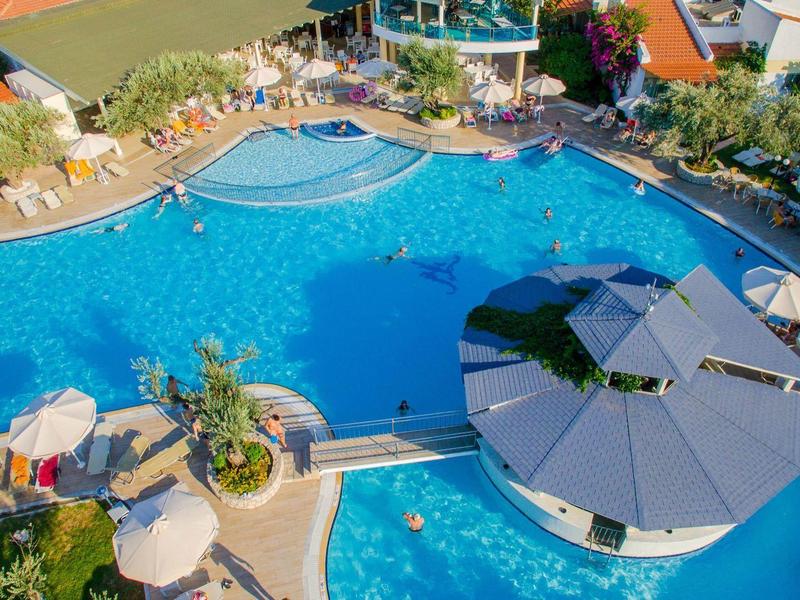 Duży basen na zewnątrz z barem swim-up i parasolami otoczony leżakami i zielenią.